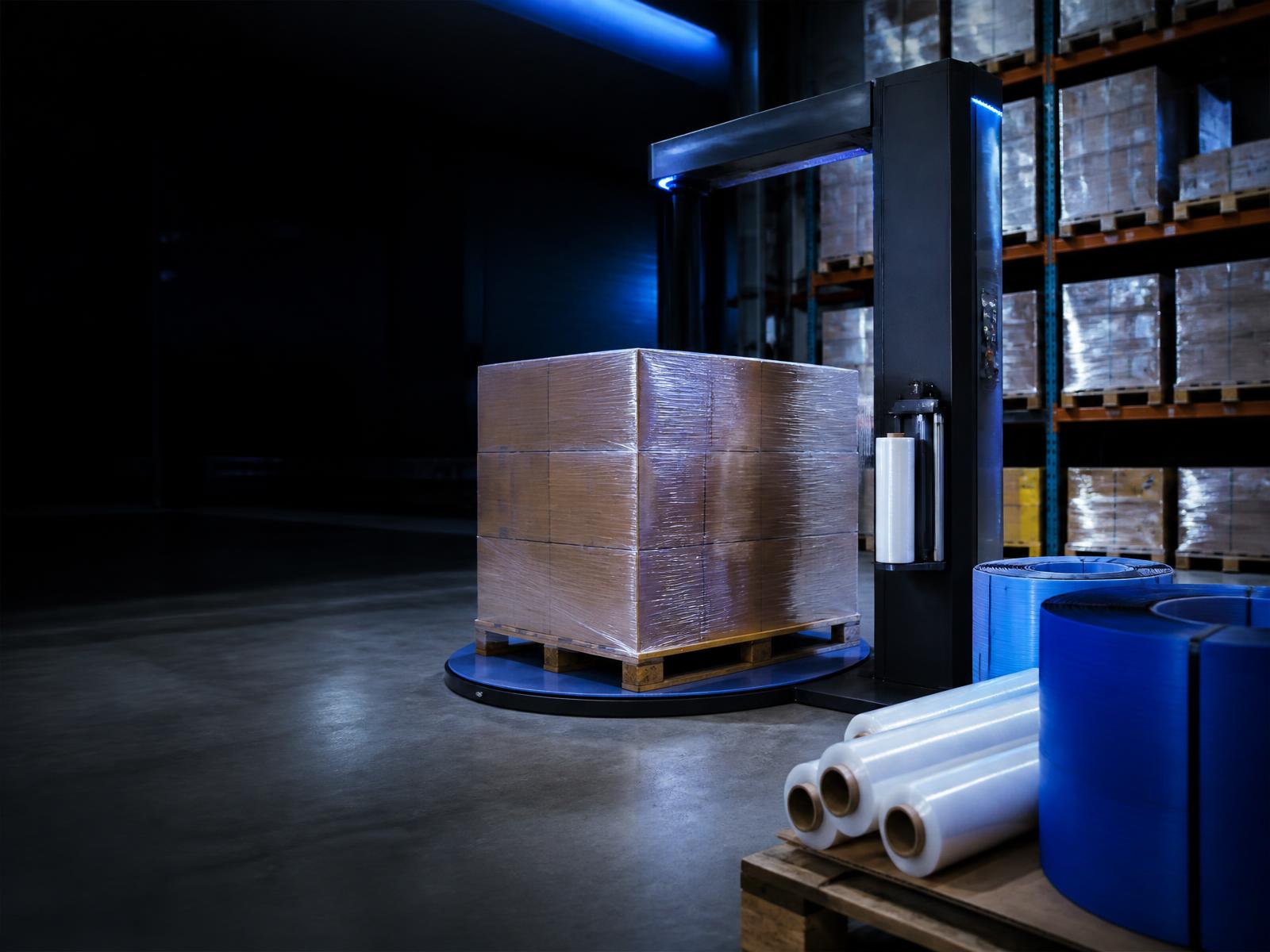 Pallet of cartons being stretch wrapped beside stretch film and strapping coils in a distribution warehouse
